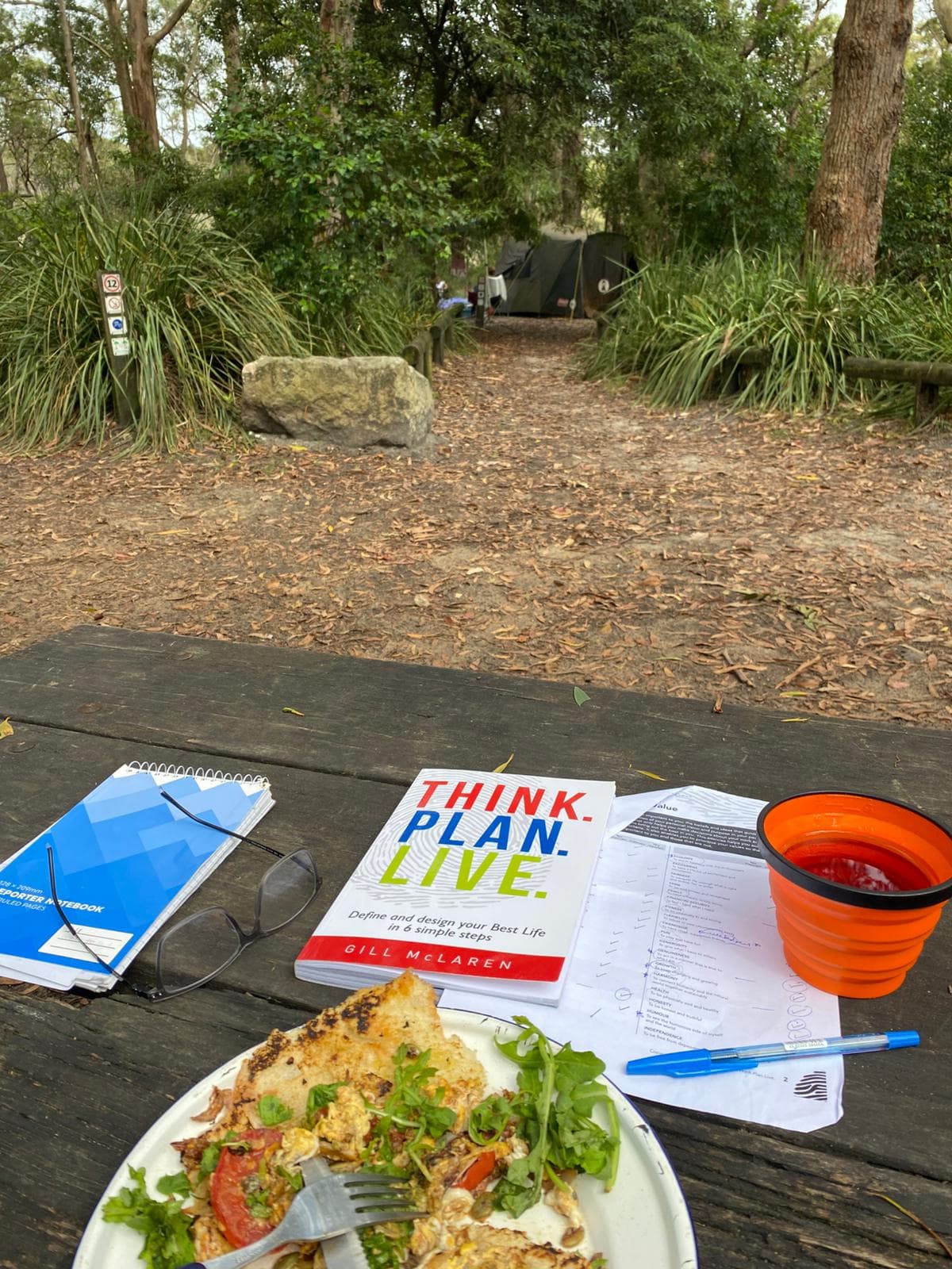 A picnic table with the Think.Plan.Live. book, a workbook and notes, set in a garden clearing.
